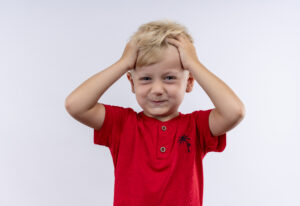 an adorable little blonde boy in red t-shirt holding hair with fingers while looking at the camera on a white background