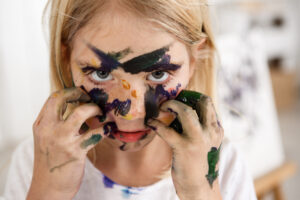 Close up portrait of beautiful little European girl with blond hair and freckles looking at the camera with her big eyes and touching her face with hands in paint, leaving colourful spots on face.