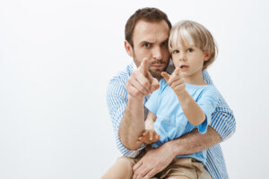 Studio shot of funny caring european father, holding and hugging cute little son with vitiligo, pointing at camera, frowning and making faces, being serious while entertaining child over grey wall