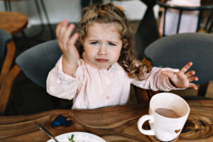 Funny little girl is sitting at a table in a restaurant, holding up hand and looking at camera. She is sitting with cacao and dessert and playing while dinner